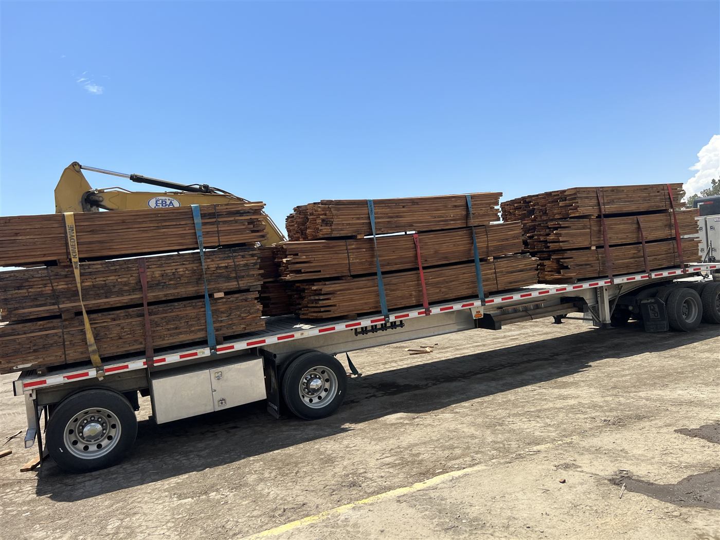 Flatbed trailer stacked with salvaged lumber being loaded by excavator